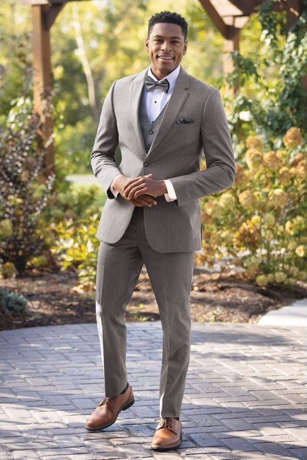 Reeves Tuxedo Rental (Medium Grey Performance Wedding Suit) - Reeves 1928 Man wearing a light gray suit, white shirt, vest and bow tie stands outdoors in a garden setting with sunshine and greenery.