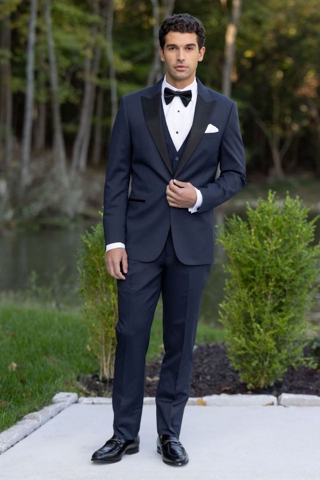 Reeves Tuxedo Rental (Navy Performance Stretch Tuxedo) - Reeves 1928 Man in a navy tuxedo with black satin lapels, white shirt, black bow tie, and pocket square standing on a garden path outdoors.