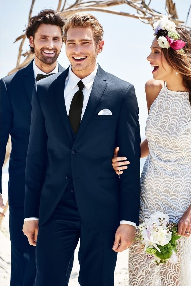 Reeves Tuxedo Rental (Navy Sterling Wedding Suit) - Reeves 1928 Wedding party on a sunny beach: groom in a navy suit walking with the bride in a white lace dress, carrying a bouquet, with a groomsman in the background.