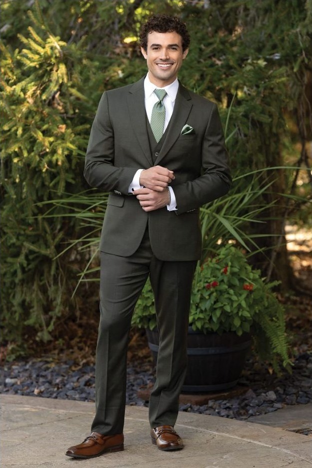 Reeves Tuxedo Rental (Olive Stretch Suit) - Reeves 1928 Man in a dark green suit with white shirt and green tie, standing outdoors and adjusting his cuffs, brown dress shoes visible.