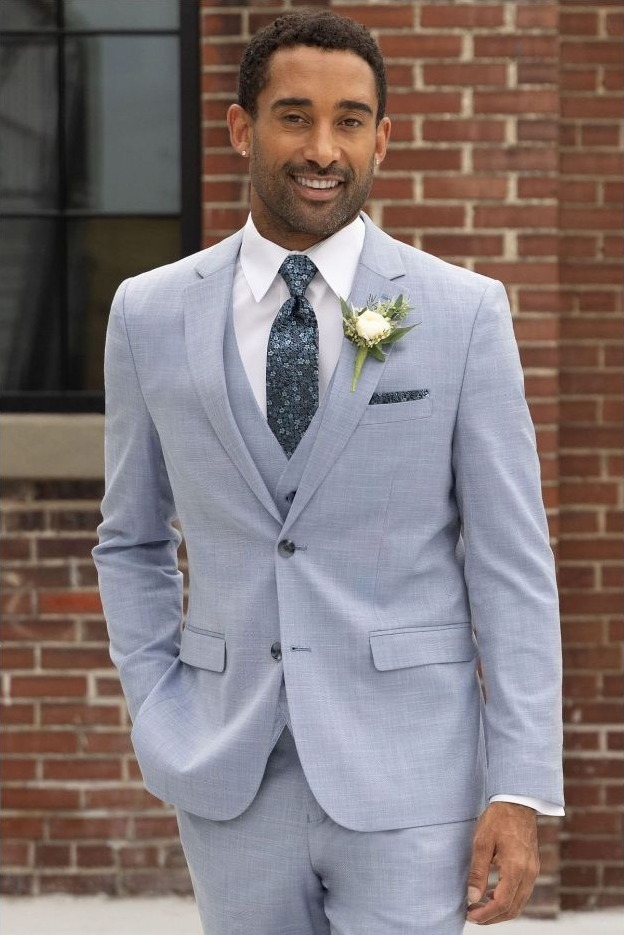 Reeves Tuxedo Rental (Oxford Blue Stretch Suit) - Reeves 1928 Man in a light gray suit with boutonniere, white shirt, and patterned tie standing outside a brick building, smiling.