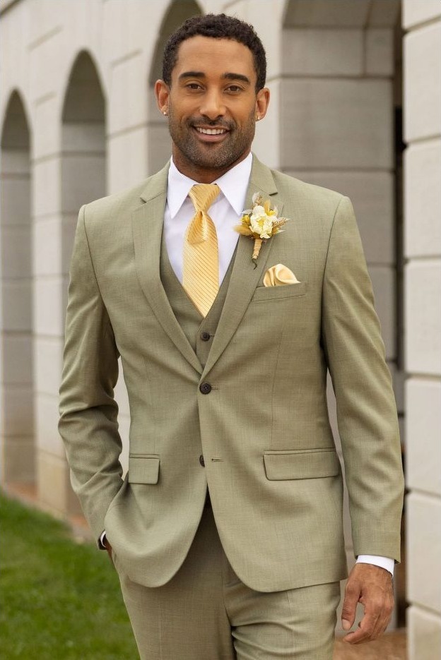 Reeves Tuxedo Rental (Sage Stretch Suit) - Reeves 1928 Smiling man in a light olive suit with a yellow tie and boutonniere, standing outdoors by a stone building.