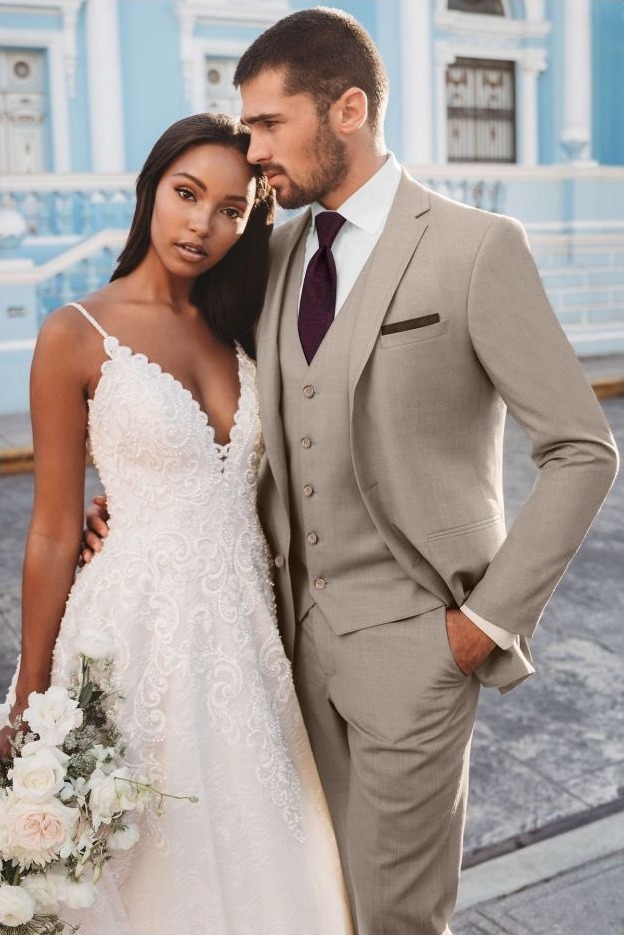 Reeves Tuxedo Rental (Sand Brunswick Suit) - Reeves 1928 Bride in a white lace wedding gown holds a bouquet beside a groom in a beige suit outdoors.