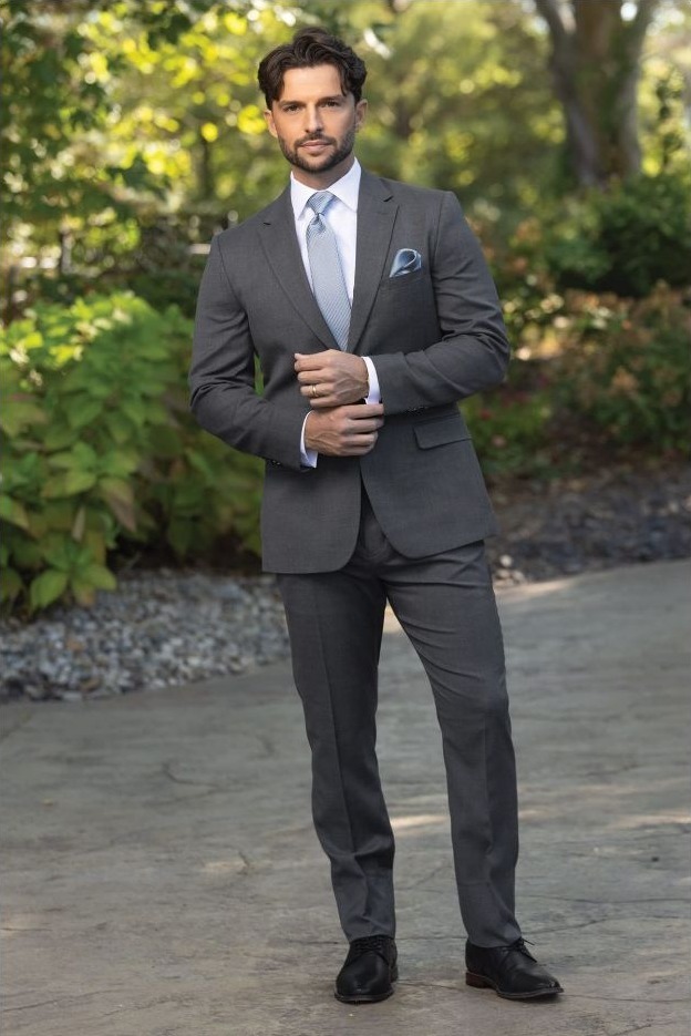 Reeves Tuxedo Rental (Slate Grey Stretch Suit) - Reeves 1928 Man in a gray suit adjusting his jacket outdoors on a sunny day, with greenery in the background.