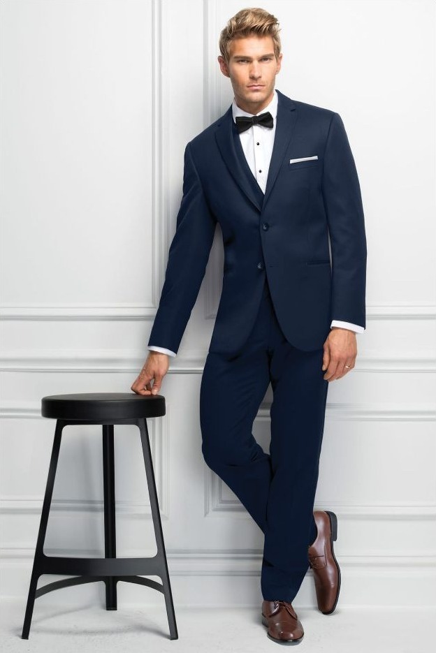 Reeves Tuxedo Rental (Slim Fit Navy Sterling Wedding Suit) - Reeves 1928 Man wearing a navy suit with a white shirt and black bow tie, leaning against a white wall beside a black stool.