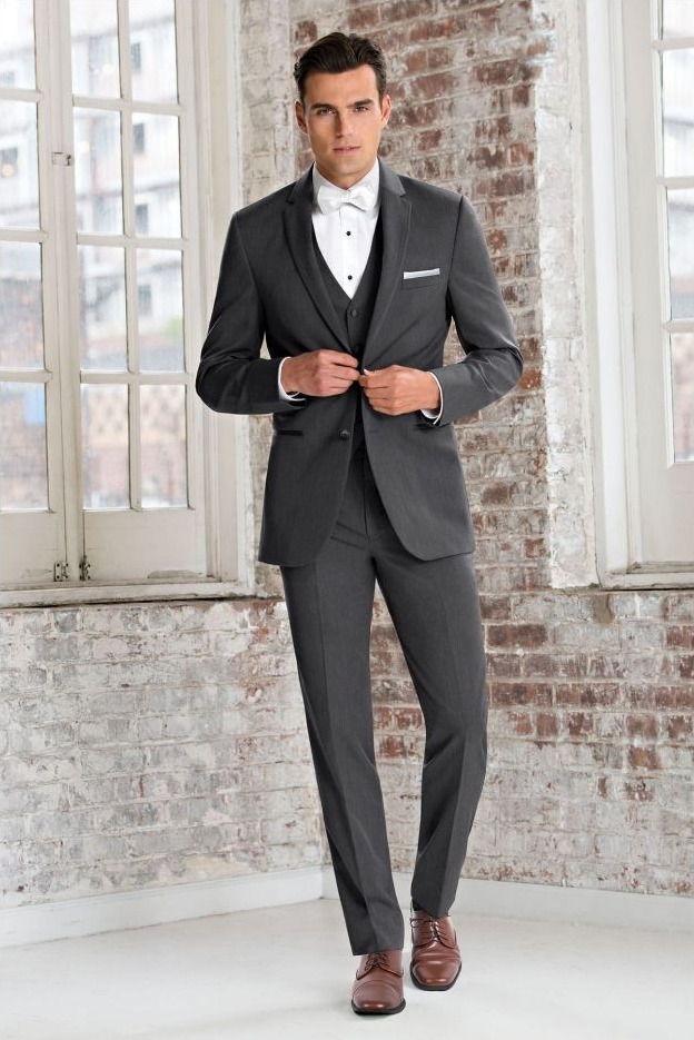 Reeves Tuxedo Rental (Slim Fit Steel Grey Sterling Wedding Suit) - Reeves 1928 Man in a dark grey suit with a white bow tie buttons his blazer, standing in a brick-walled room with large windows.