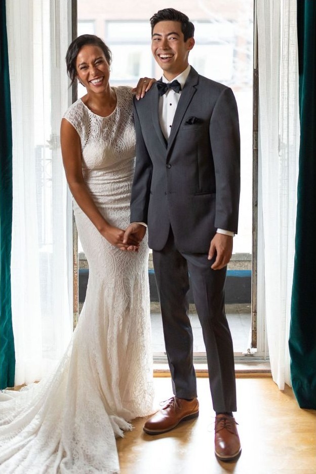 Reeves Tuxedo Rental (Steel Grey Sterling Wedding Suit) - Reeves 1928 A smiling bride in a white lace gown stands beside a groom in a dark gray suit and bow tie, holding hands by a sunlit doorway.