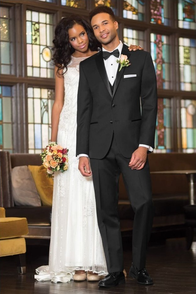 Reeves Tuxedo Rental (Super 120s Black Tuxedo) - Reeves 1928 Bride and groom posing together in a wedding venue; the bride in a white gown holding a bouquet, the groom in a black tuxedo with bow tie.