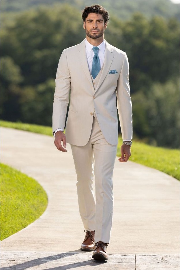 Reeves Tuxedo Rental (Tan Performance Wedding Suit) - Reeves 1928 Man in a light beige suit with white shirt and teal tie, walking along a sunlit park path, brown dress shoes and pocket square visible.