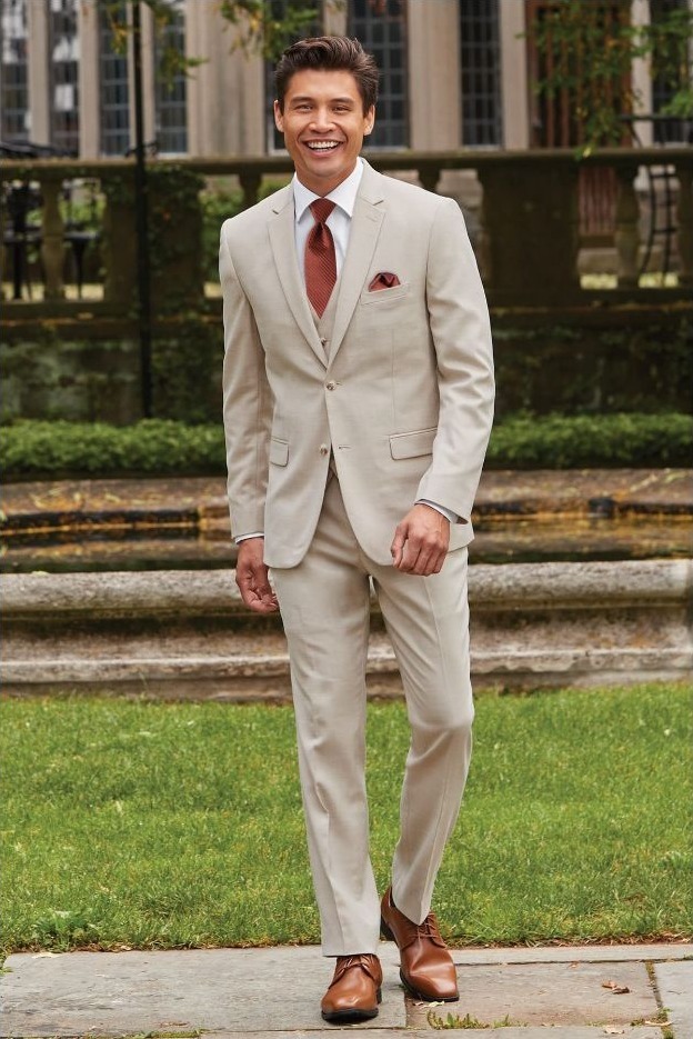 Reeves Tuxedo Rental (Tan Stretch Suit) - Reeves 1928 Man in a light beige suit walking outdoors, smiling, with brown dress shoes and a red tie in a garden setting.
