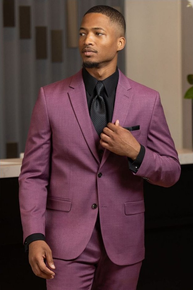 Reeves Tuxedo Rental (Violet Stretch Suit) - Reeves 1928 Man in a mauve suit with black shirt and tie, standing confidently indoors, adjusting his lapel.