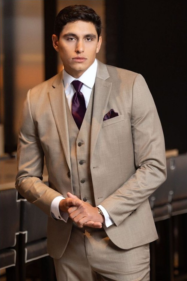 Reeves Tuxedo Rental (Wheat Stretch Suit) - Reeves 1928 Man in a beige three-piece suit with a burgundy tie and pocket square, standing in a modern office setting.