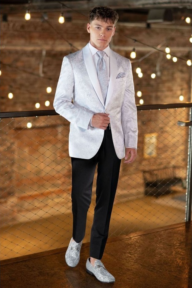 Reeves Tuxedo Rental (WhiteSilver Paisley Aries Tuxedo) - Reeves 1928 Young man wearing a light silver tuxedo jacket, matching tie, black pants, and glittery silver loafers in a warmly lit space with string lights.