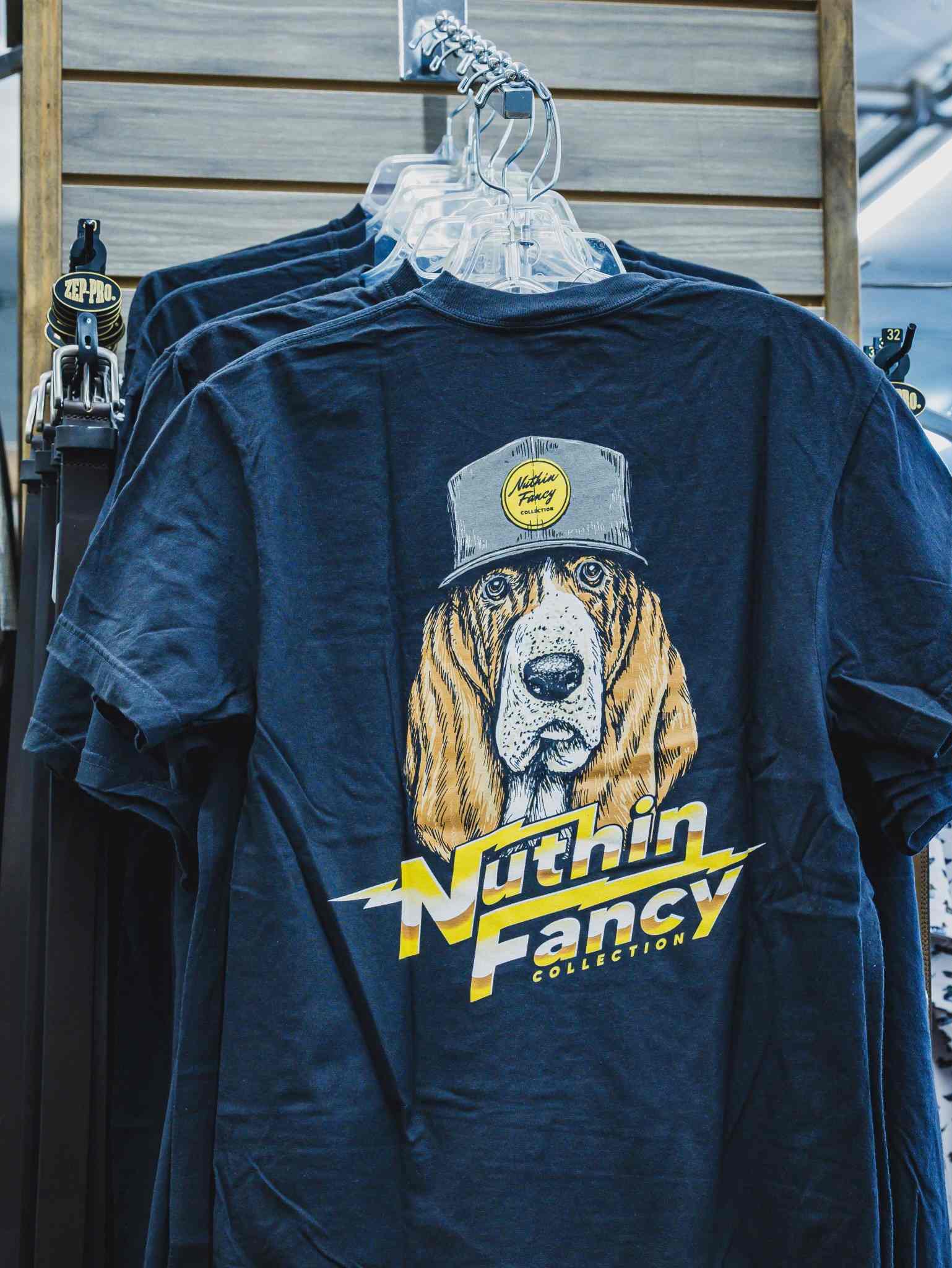 Blue T-shirt on clear hangers with a cartoon dog wearing a cap and bold yellow lettering graphic on the chest area