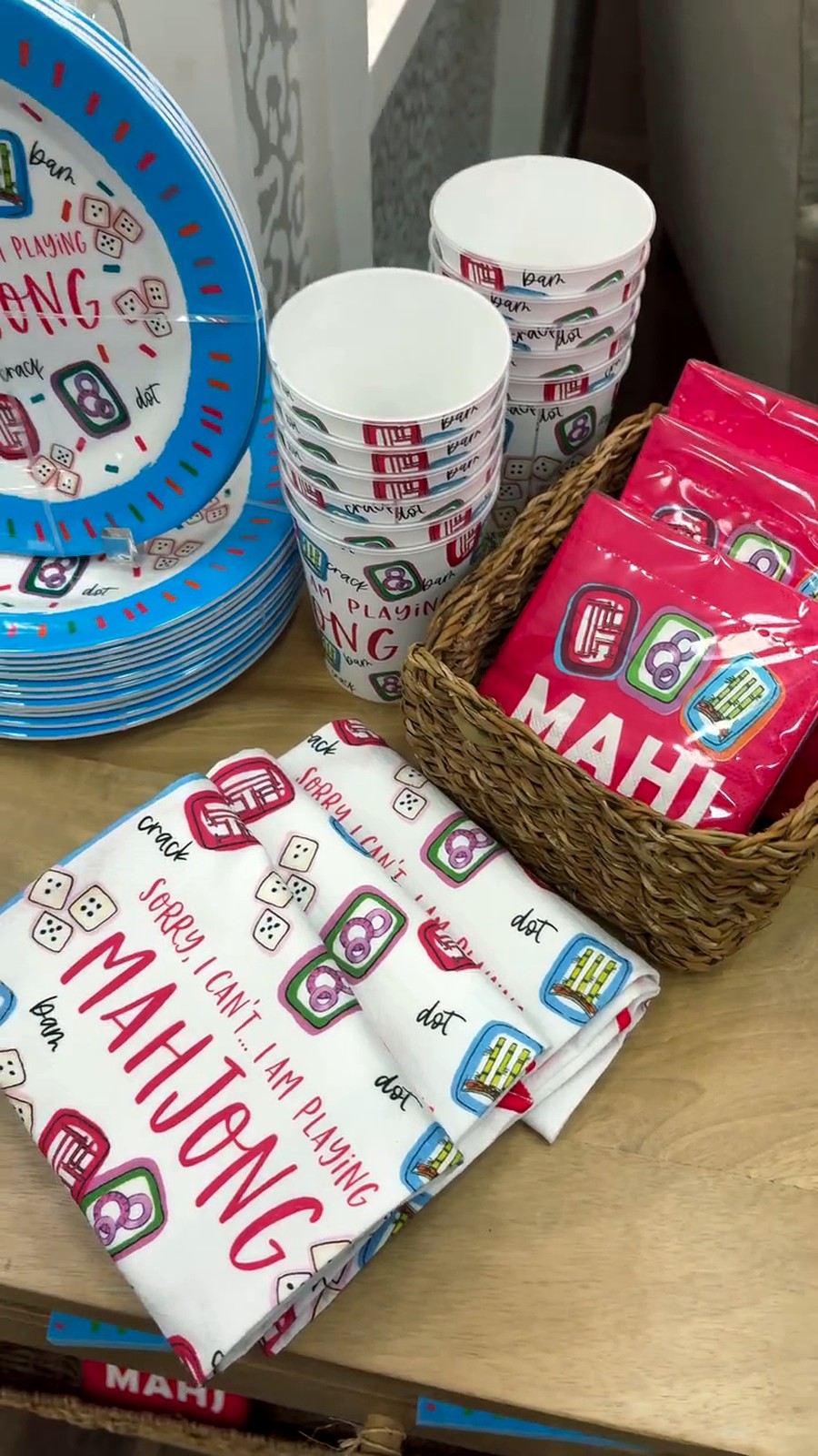 Colorful party supplies featuring Mah Jong-themed plates, cups, napkins, and red packets arranged in a basket and on a table.