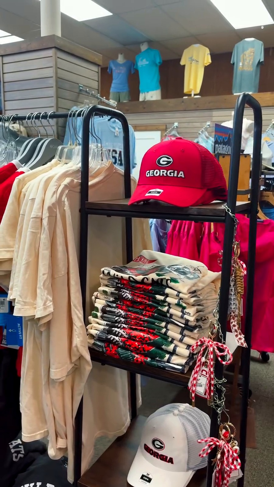 Red Georgia cap with 'GEORGIA' text on display rack in a clothing store, with stacks of t-shirts nearby.