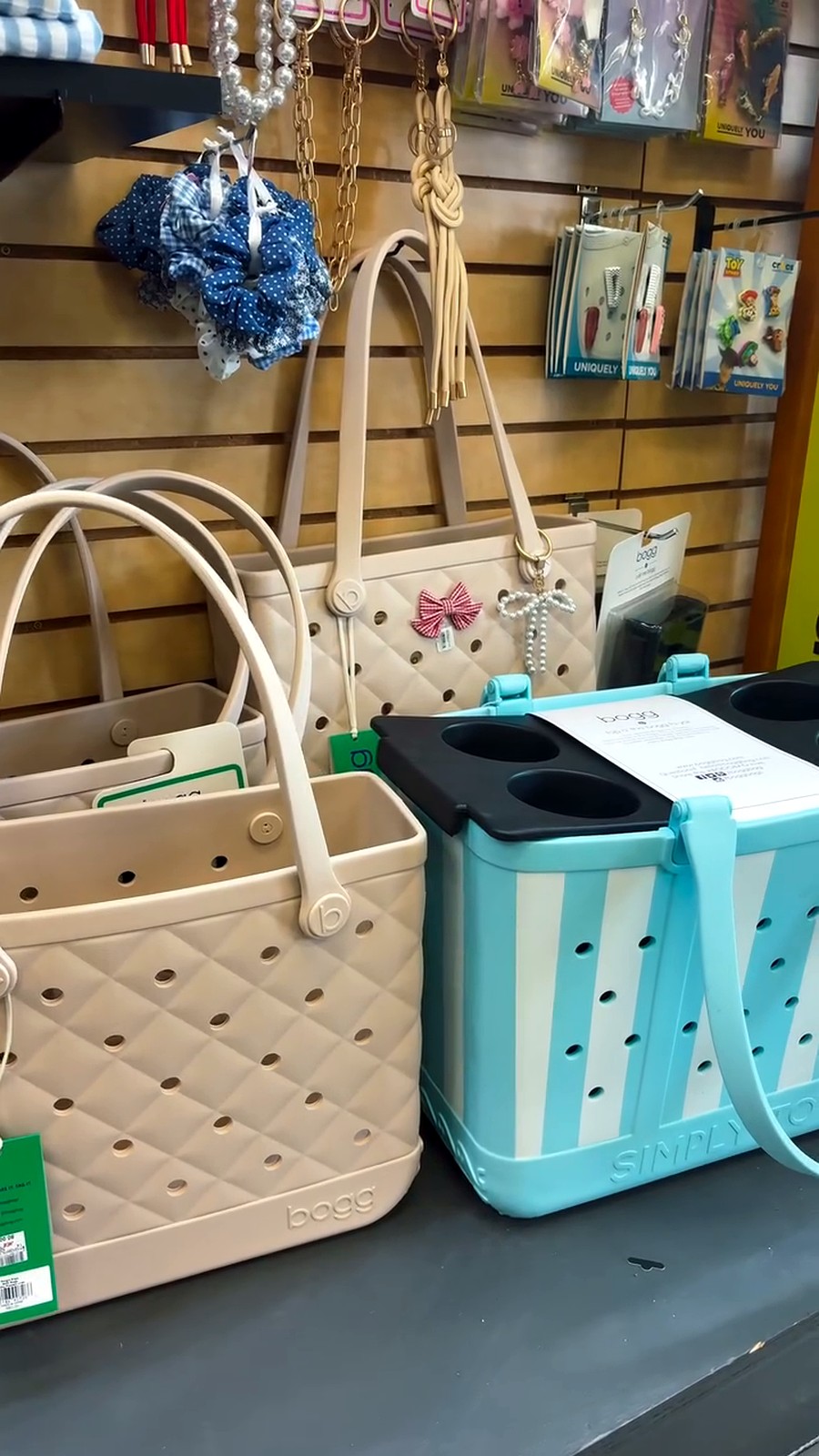 Display of handbags and accessories on a store rack: a beige quilted tote with a pink bow charm beside a blue striped tote, with jewelry and packaging hanging above.