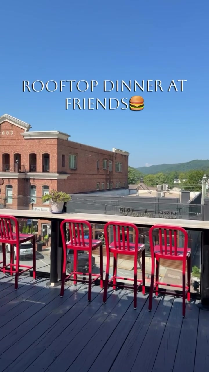 Rooftop deck with bright pink chairs along a glass railing, city rooftops below and green hills in the distance under a clear blue sky, overlaid with 'ROOFTOP DINNER AT FRIENDS 🍔'.