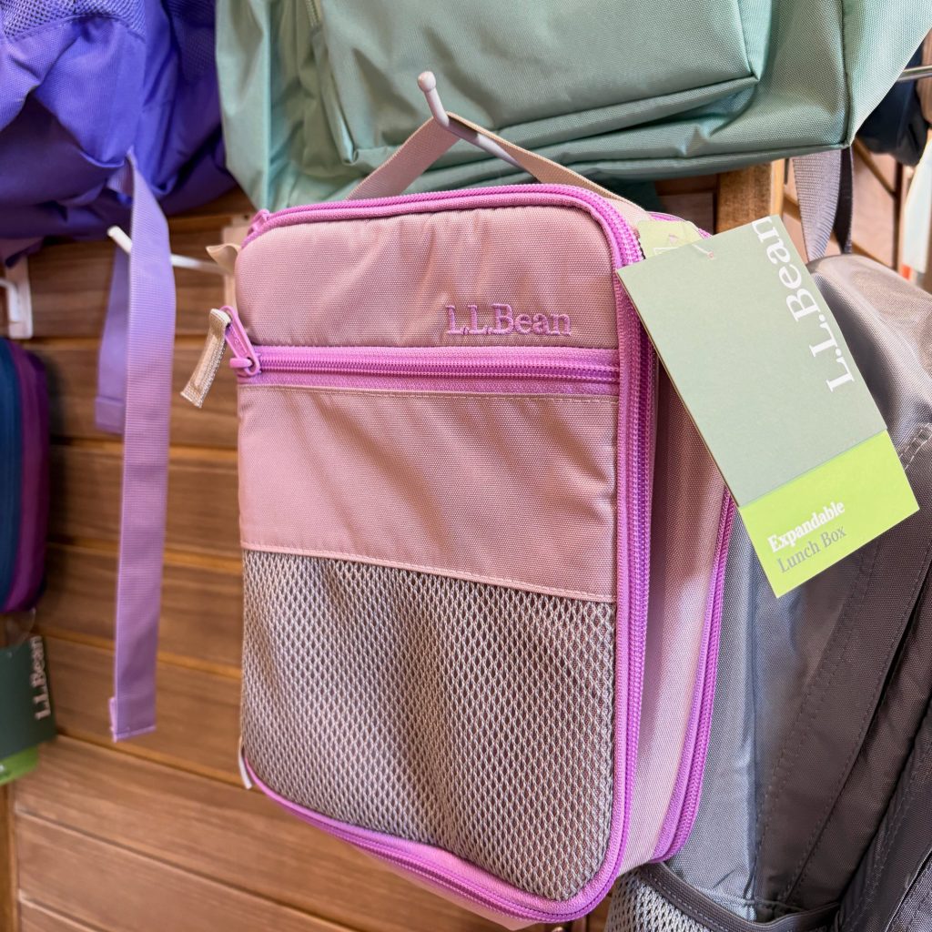 Pink LL Bean backpack with pink zipper, hanging tag visible, among green and purple backpacks on a shelf item.