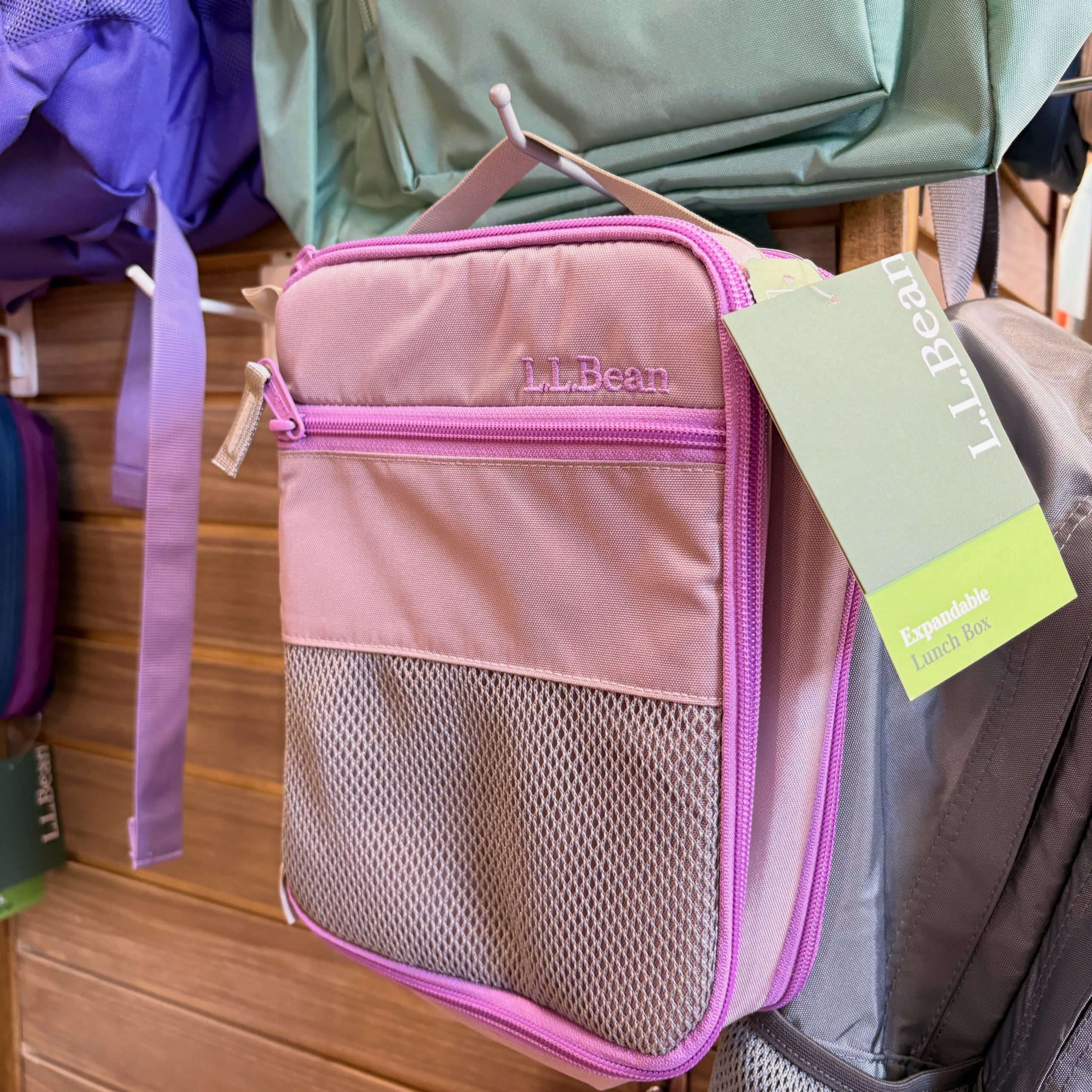 Pink LL Bean backpack with pink zipper, hanging tag visible, among green and purple backpacks on a shelf item.