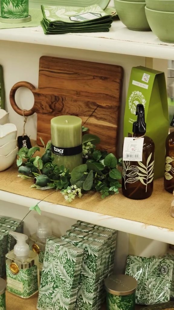 Green shelf display with a wood cutting board, a green candle wrapped in black label, and a brown glass bottle with a white leaf design; decorative greenery surrounds them on a beige mat.