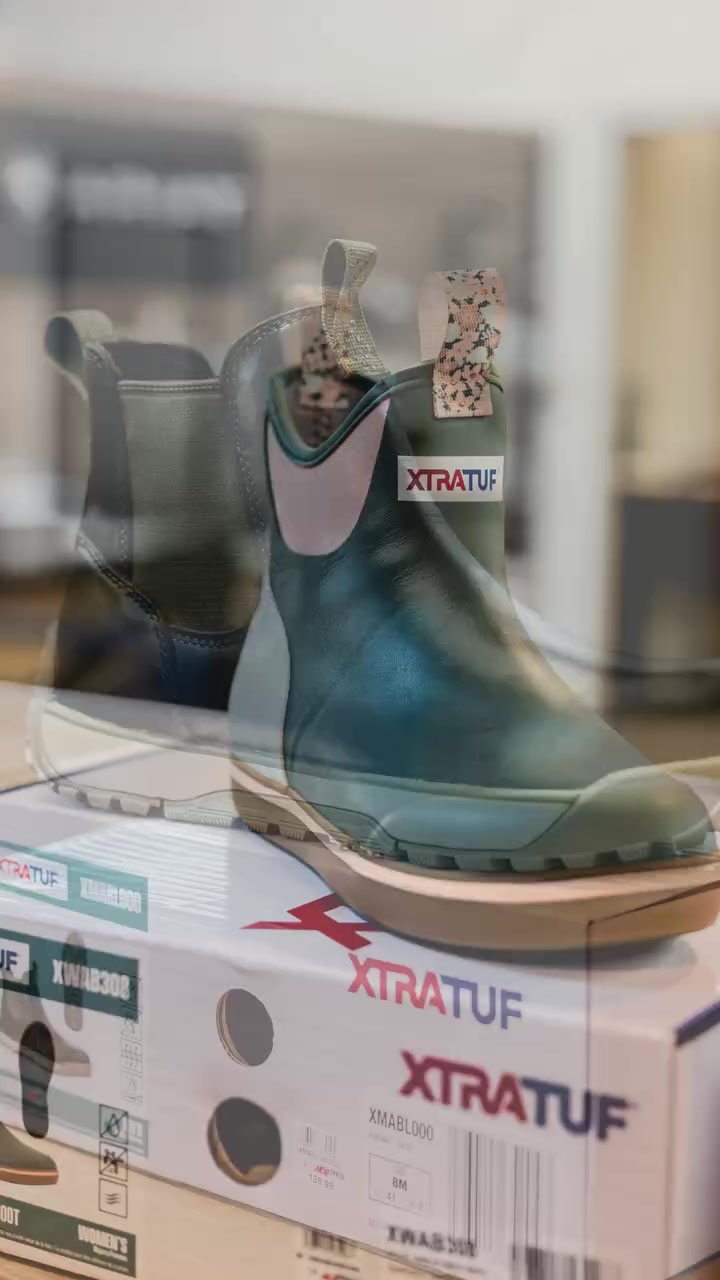 Dark teal XTRATUF waterproof boot with pink pull tab on a product display box in a store window reflection
